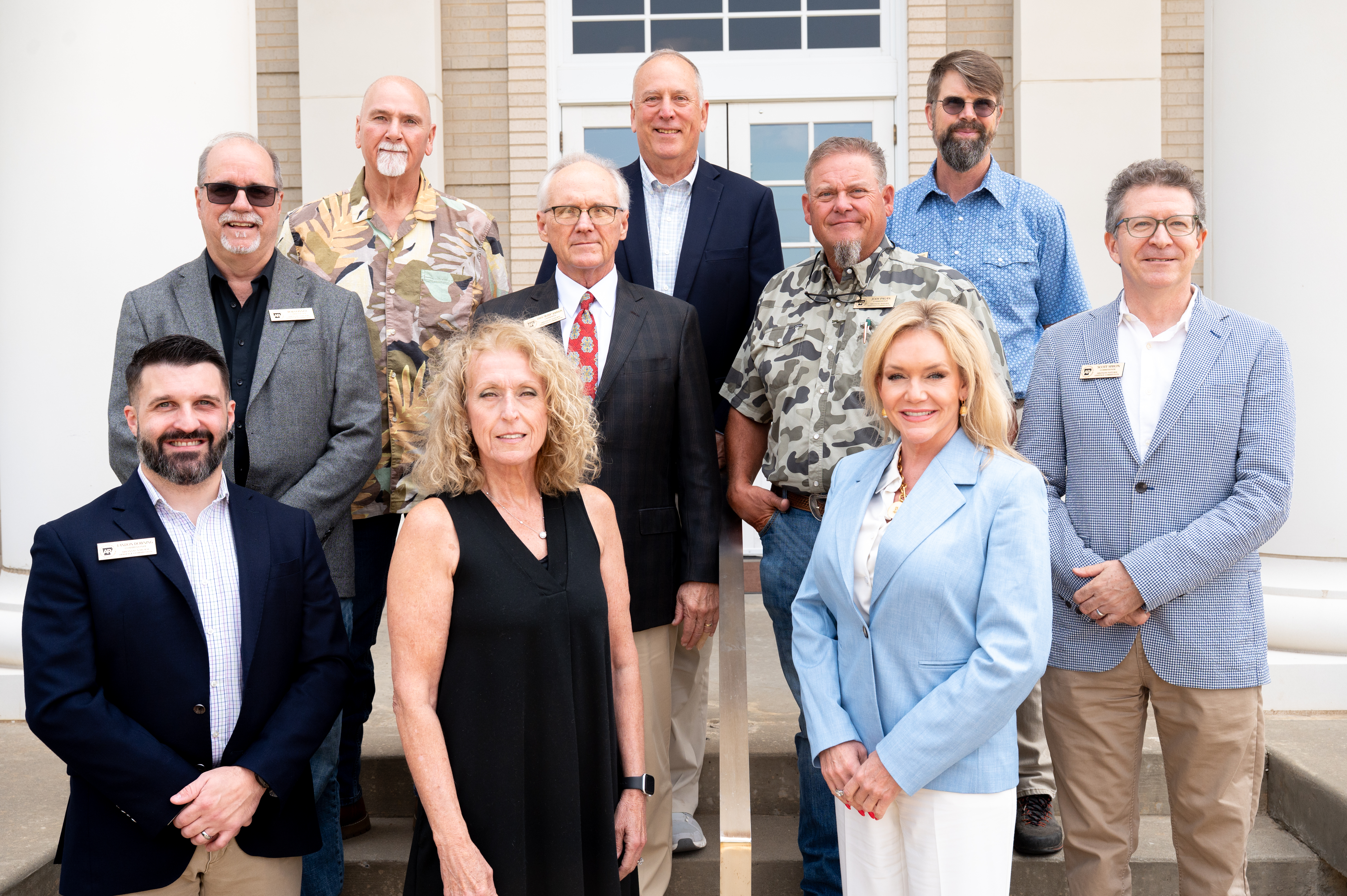 Group of people in business casual clothing posing for a photo on steps in front of a cream brick building.