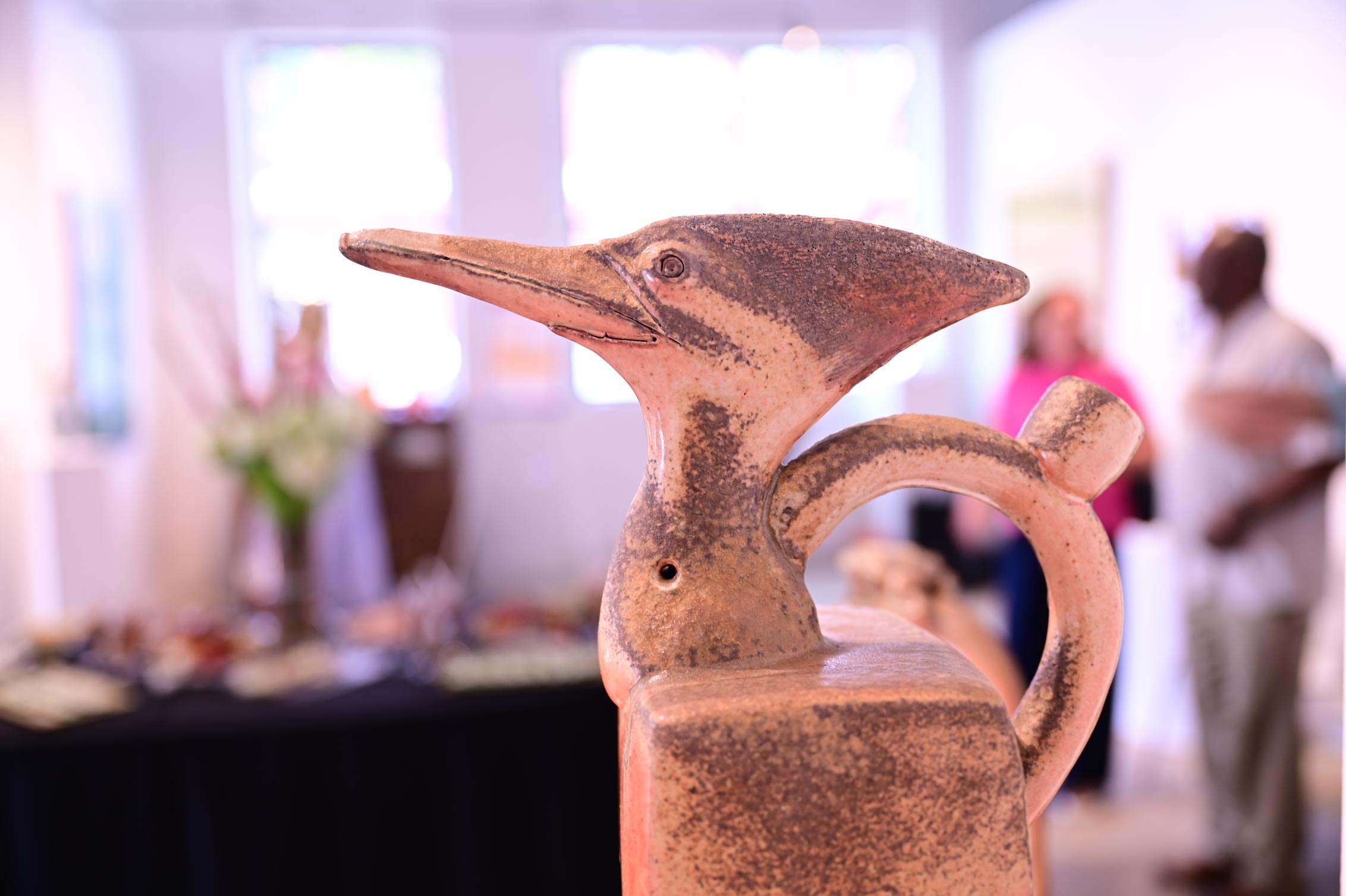 A clay object shaped as a birds head and face with a handle on its back all on an organically shaped cube block.