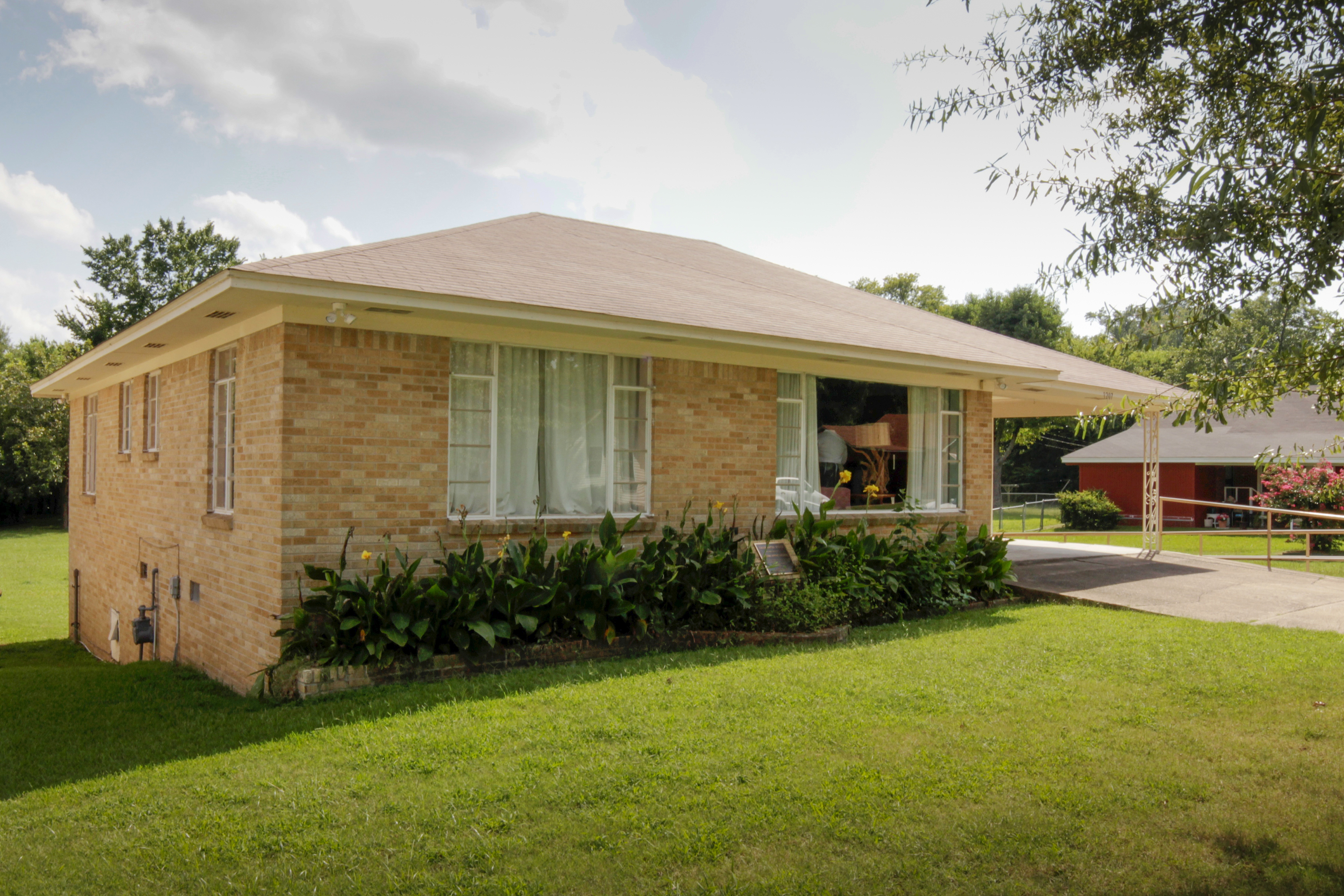 Front view of the Daisy Bates Home. Two large front windows with white curtains surrounded by yellow-gold bricks and foilage lining the front wall.