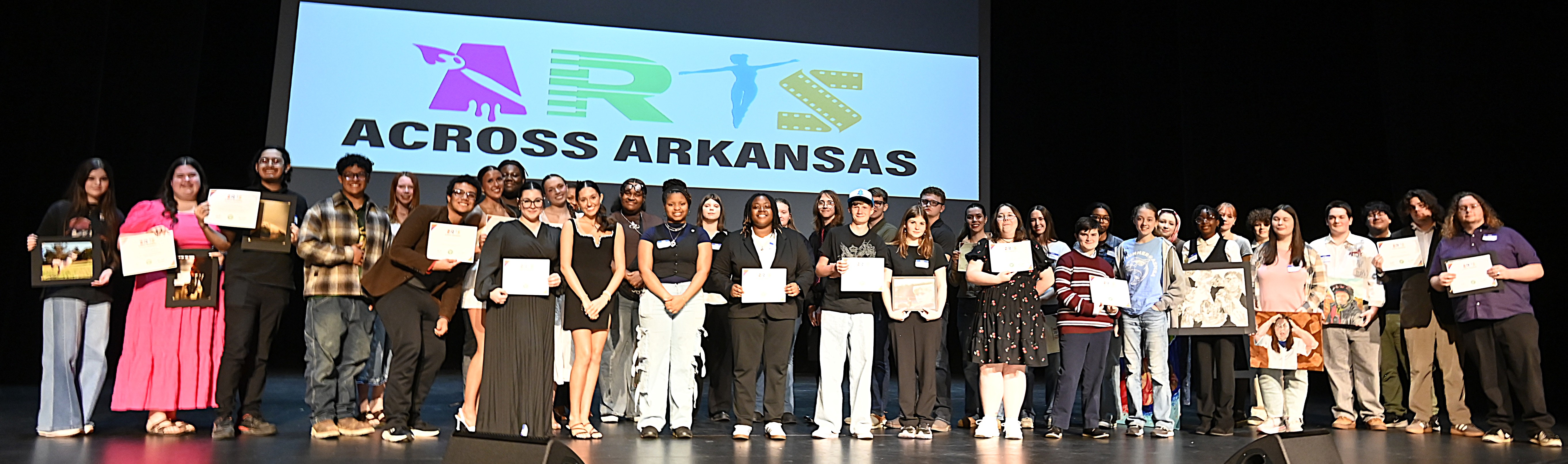 About 54 high schoolers pose on stage in front of the Arts Across Arkansas logo.