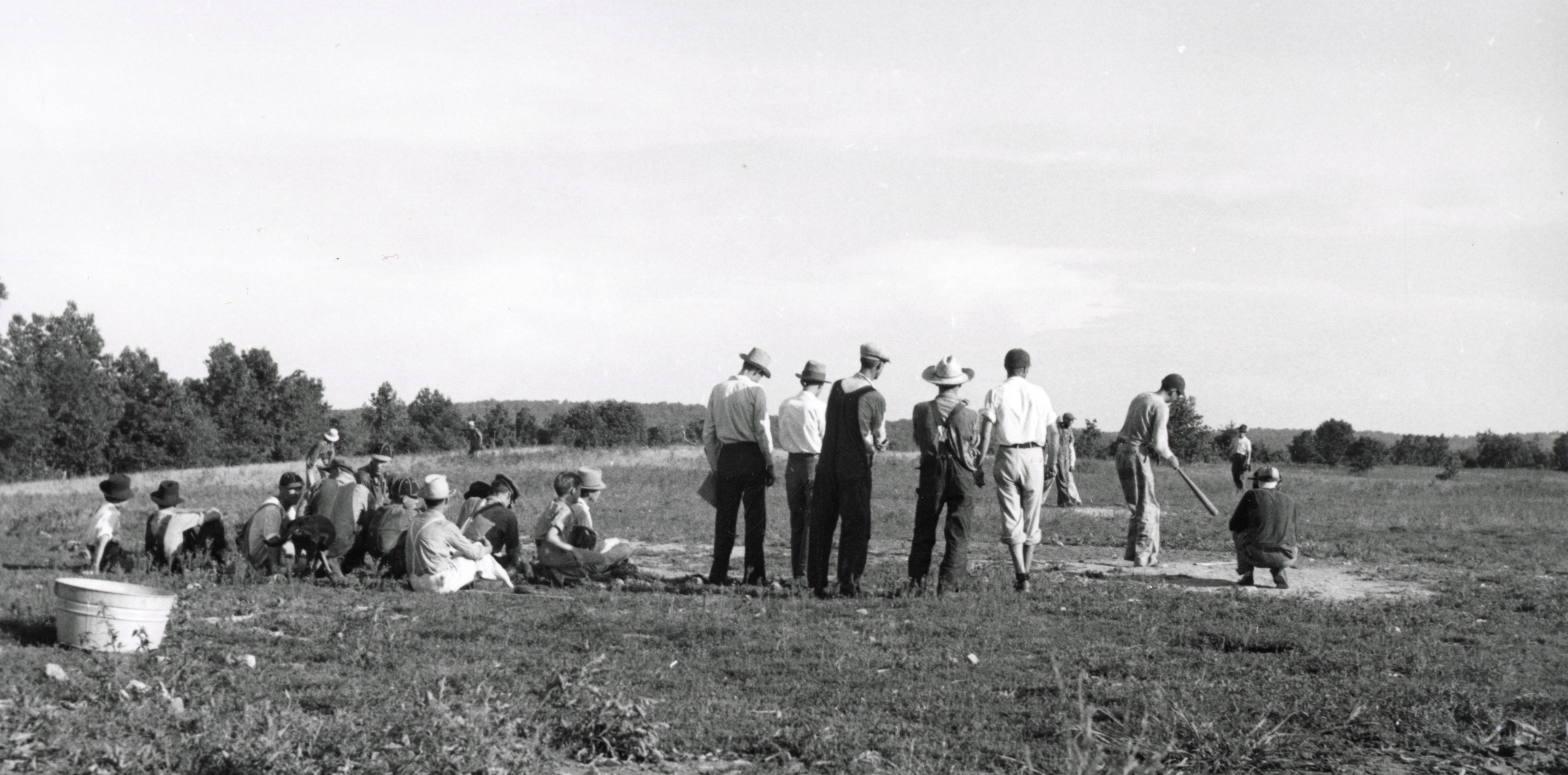 A group of men and boys with some playing baseball while others sit on the grass and watch.