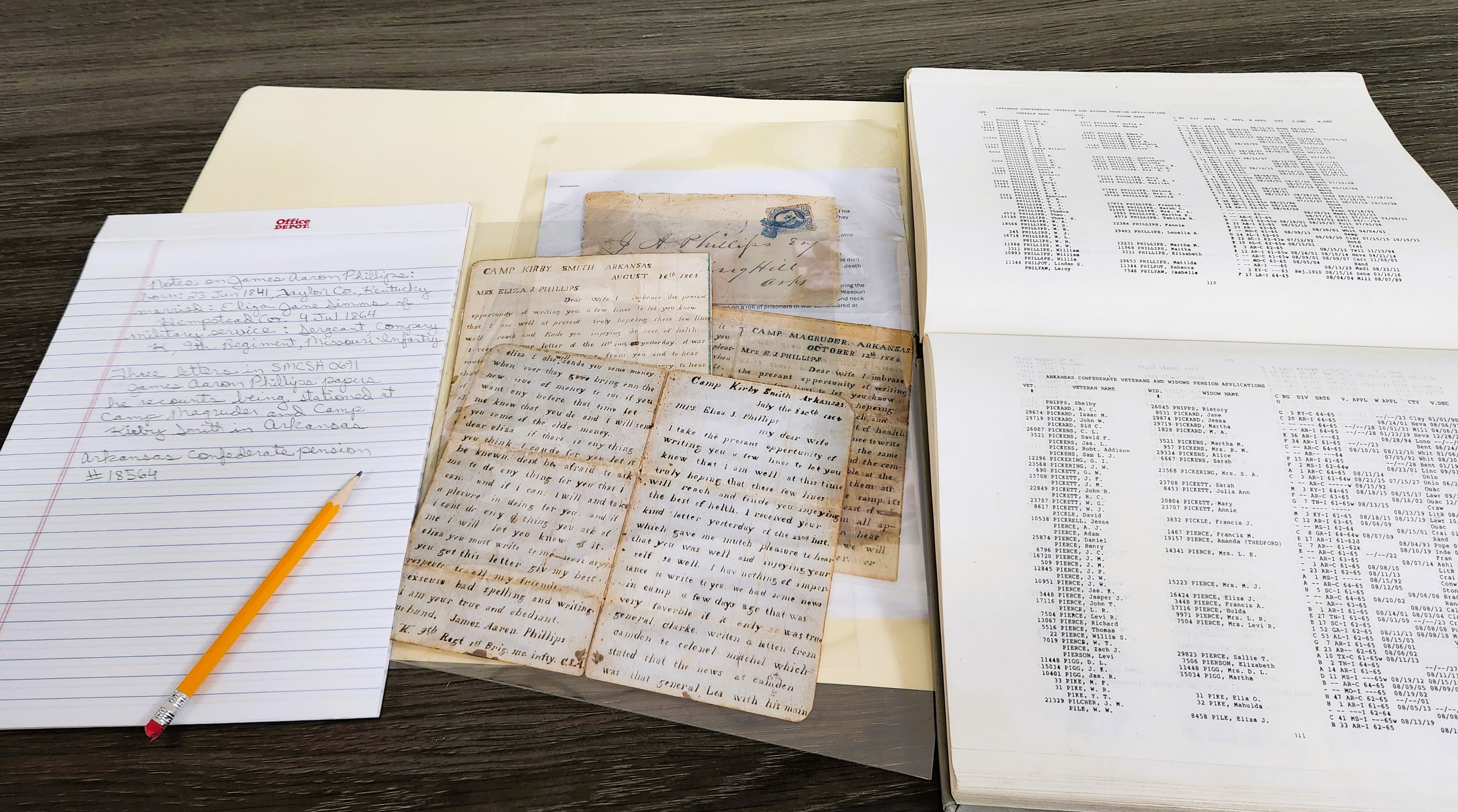 A spread of old documents and ledgers on a table with a notepad and pencil to the left.