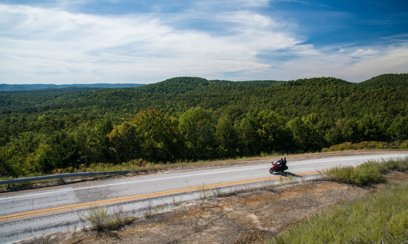 A motorcyclist rides along the road next to a hilly tree covered view.