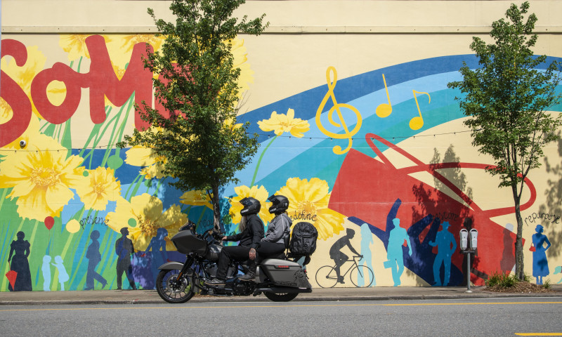 Two people ride a motorcycle on pavement next to a large colorful mural.
