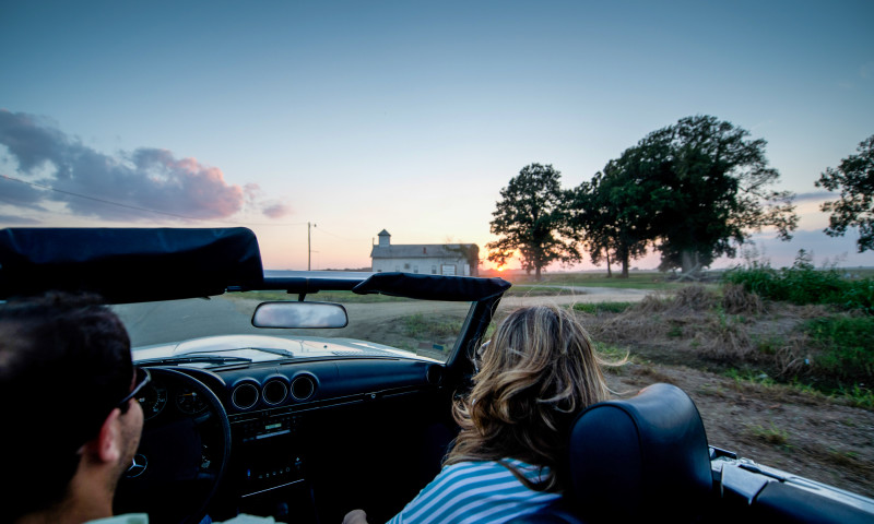 A couple driving in a convertible car along an open road with a few trees and a house in the distance at sunset.