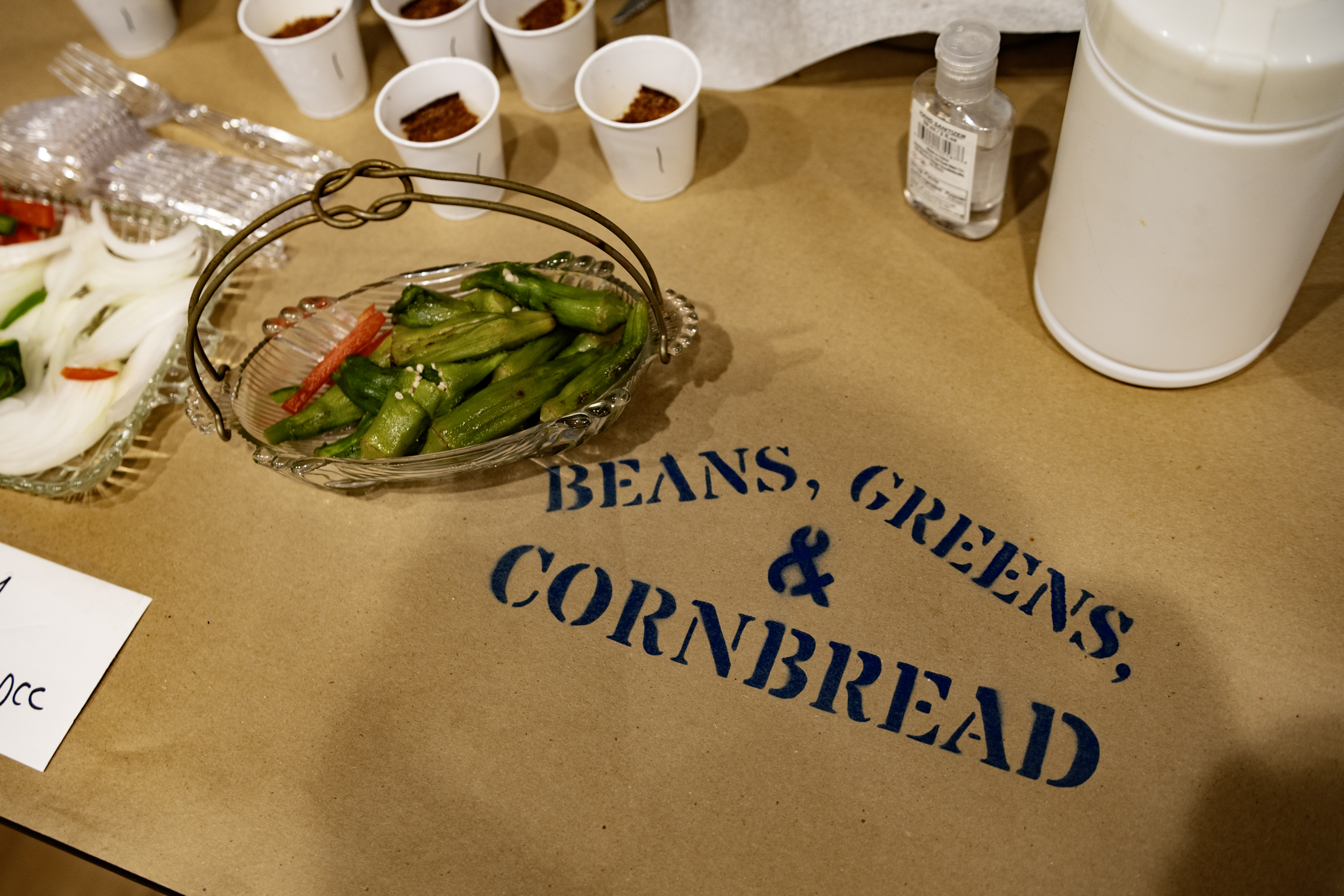 A paper table cloth with the words Beans, Greens, & Cornbread printed on it. Peppers and other food items sit along the table.