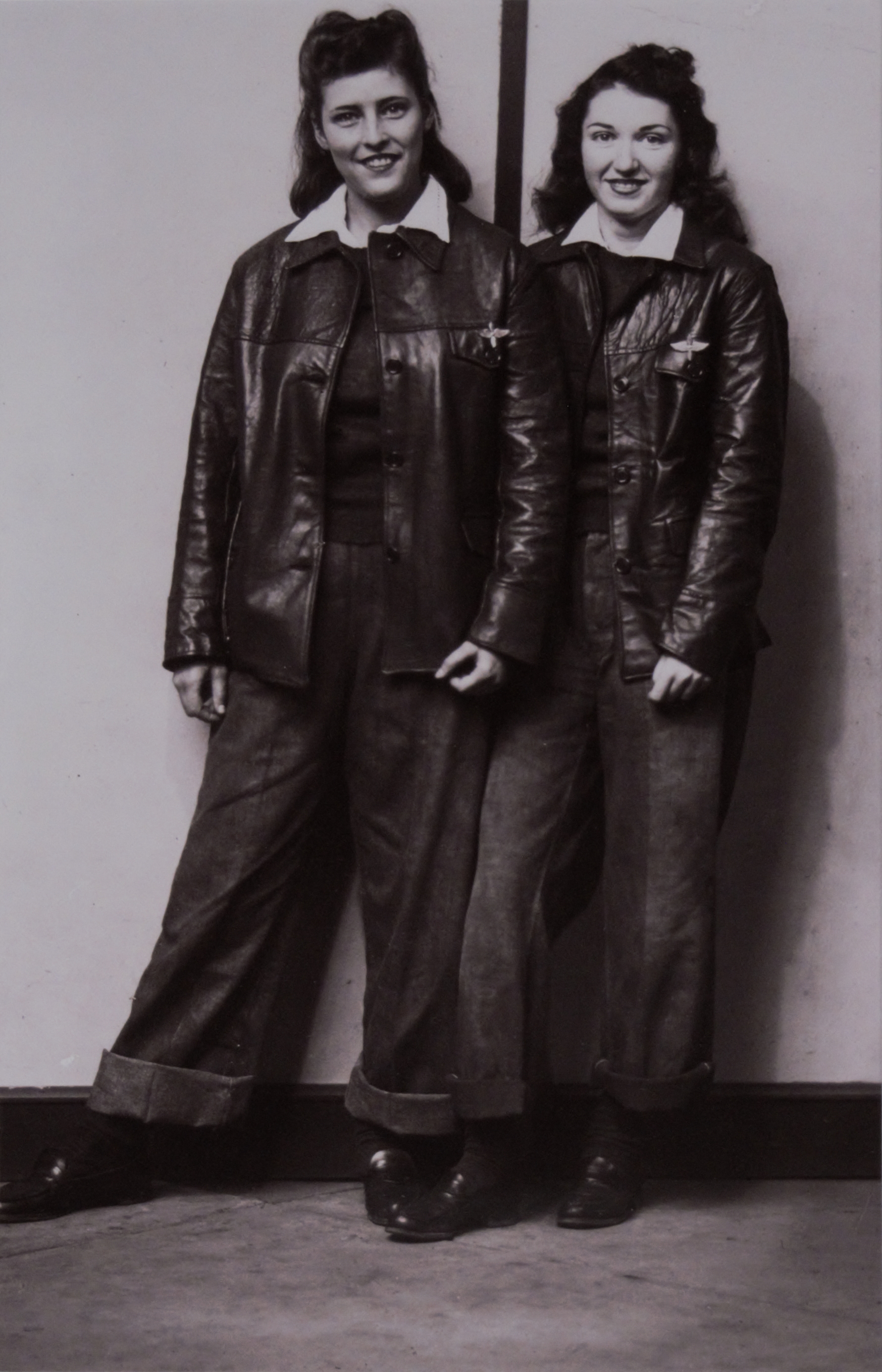 Two young ladies pose next to one another in leather jackets and cuffed jeans.