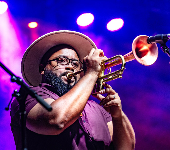 A man on stage in front of blue and purple lights playing trumpet into a microphone.