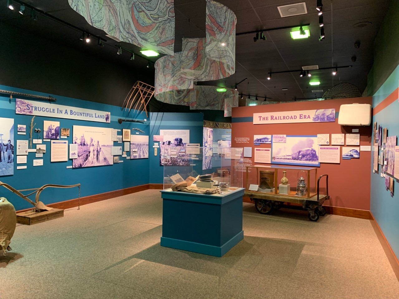 A large exhibit room with text and imagery lining blue and orange walls. Several historic objects are on the floor and in the center is a lightbox with more objects.