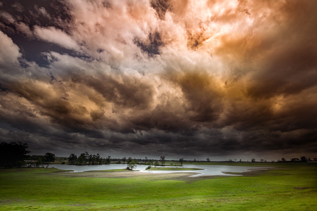 Dramatic lighting of an open scene of a cloudy sky and a green grassy field with a large pond of water in the distance.