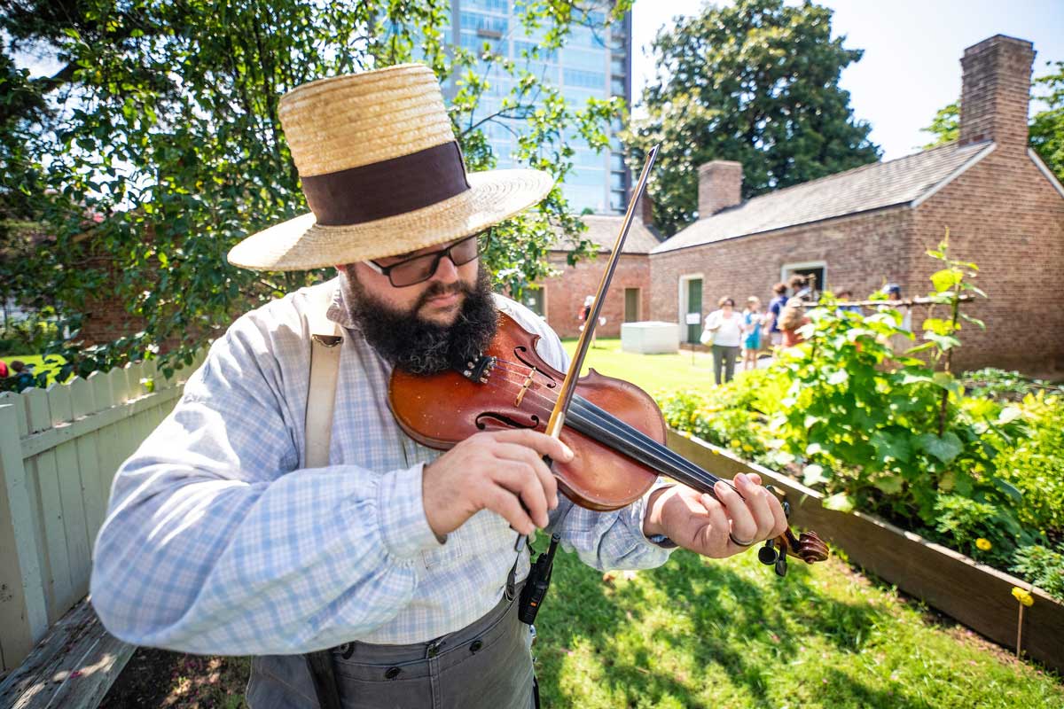 A man in suspenders and a wide brimmed straw hat fiddling on historic grounds.