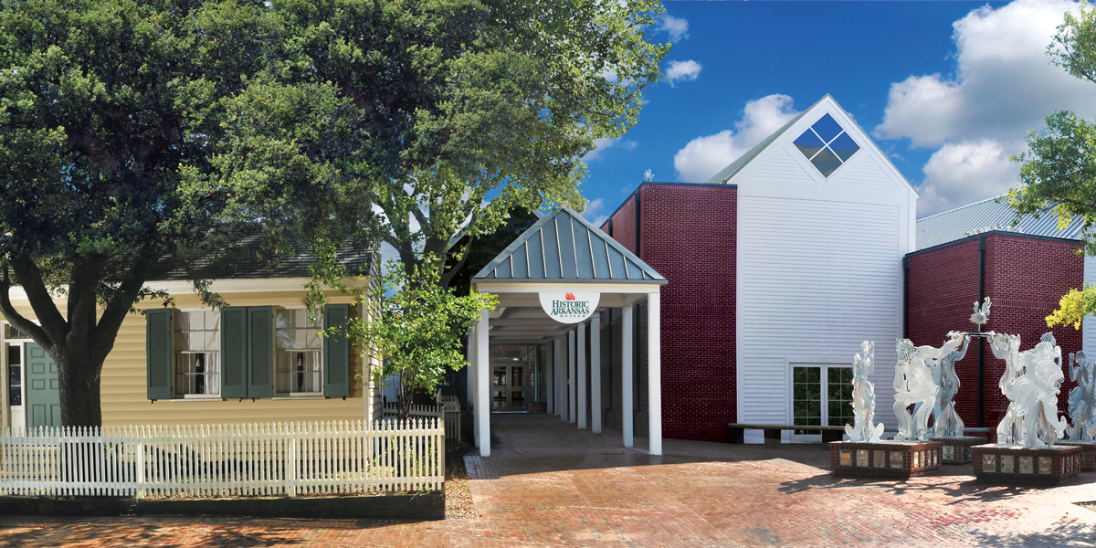 A yellow house with trees in the front to the left, a green-roofed awning with "Historic Arkansas Museum" written on it in the center, and a brick and white sided building to the right with statues in front.