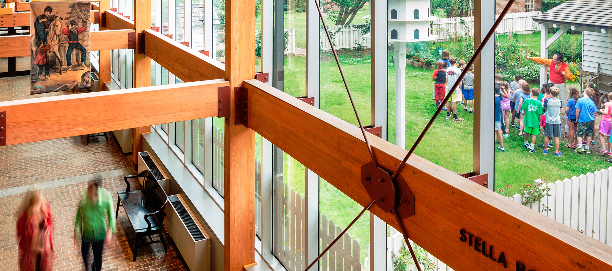 Wooden structural beams above two adults walking inside and a group of children walking outside.