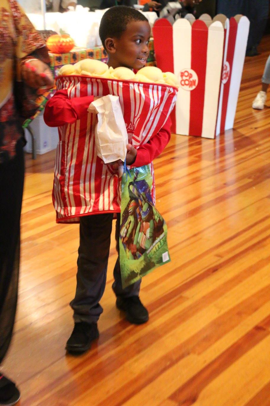 A young boy eats popcorn in a popcorn halloween costume holding a bag of halloween candy.
