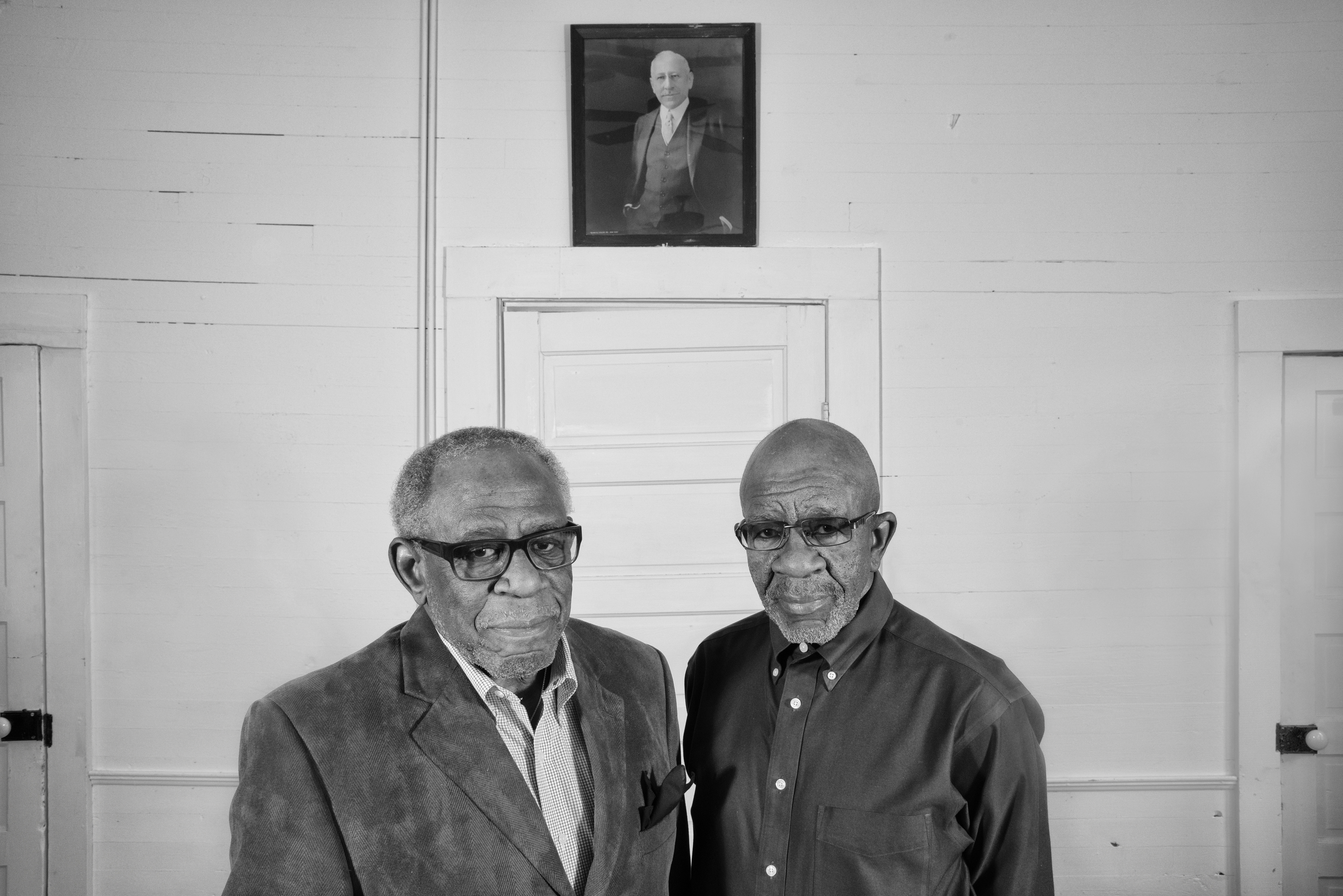 Two men posding side by side in front of a door under a picture portrait.