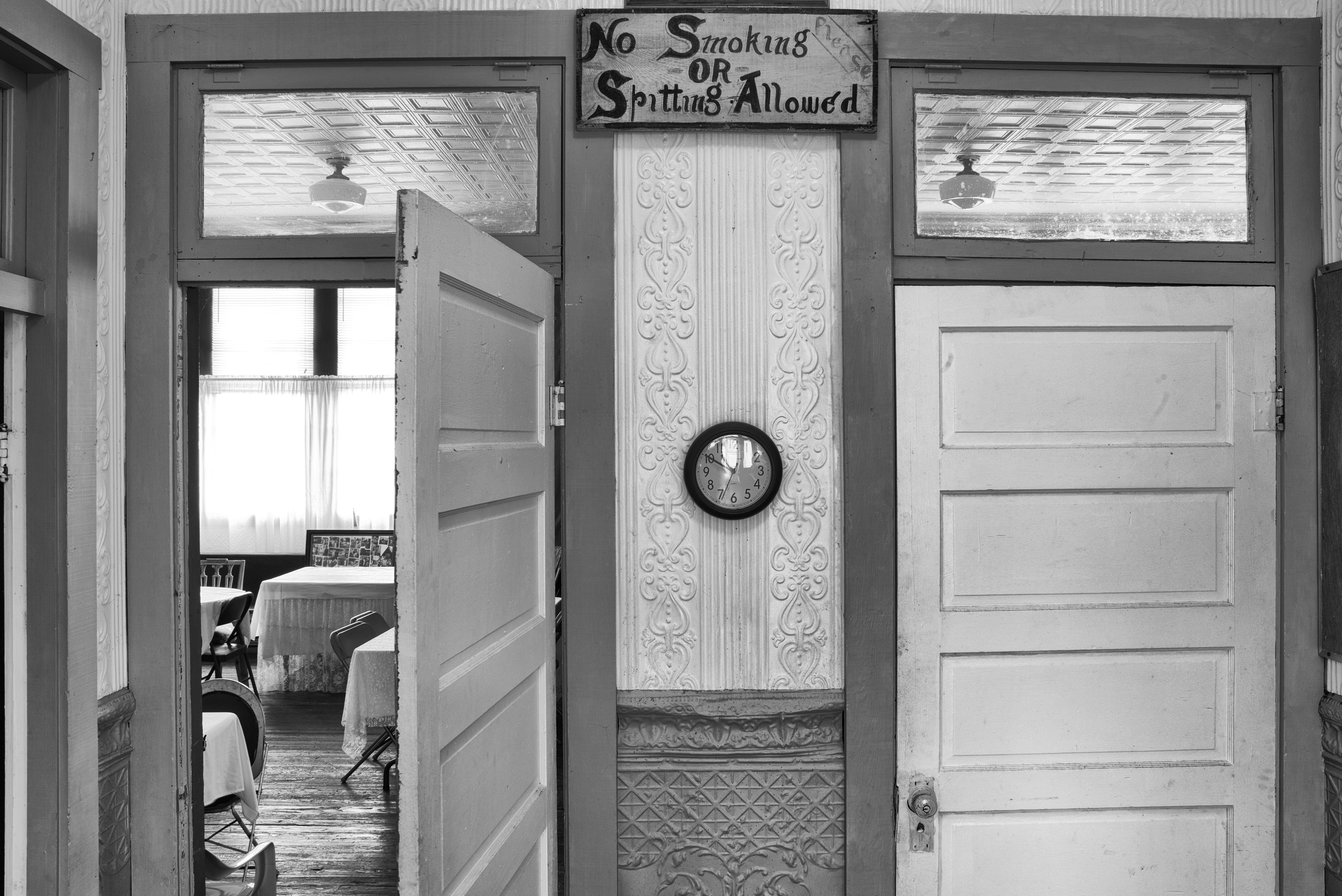 Facingawallwithtwodoorsandaclockinthecenterwiththeleftdooropenshowingtablesandchairs