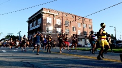 Agroupofcontestantsinathleticwearrunningpastabuildinginarace