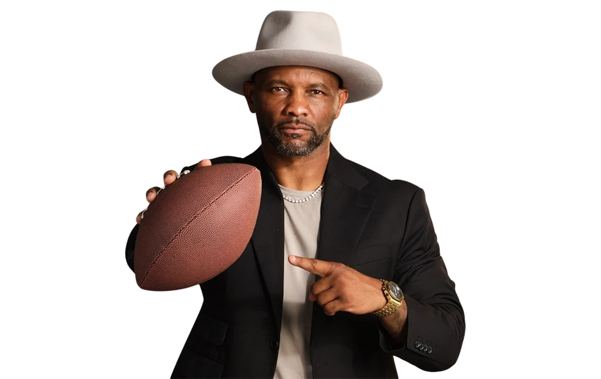 Amaninablackblazerandagrayhatholdsafootballinhisrighthandandpointstoitwithhisleft