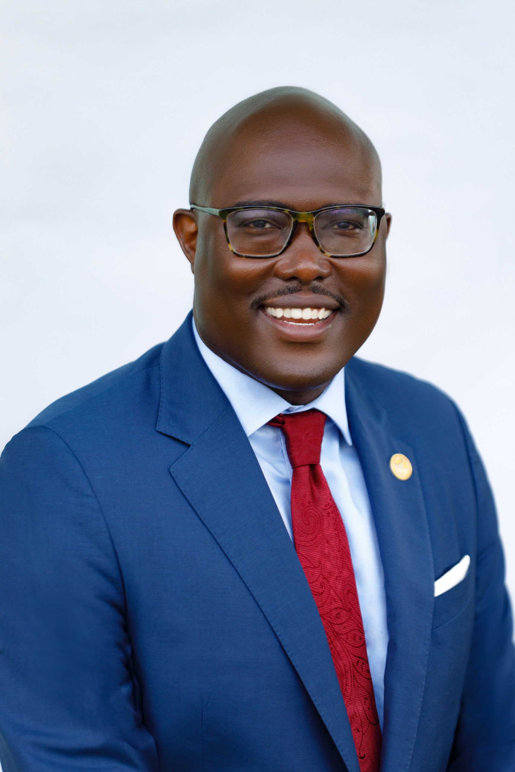 Frank Scott Jr., a man with dark colored glasses in a blue suit with a red tie smiling.