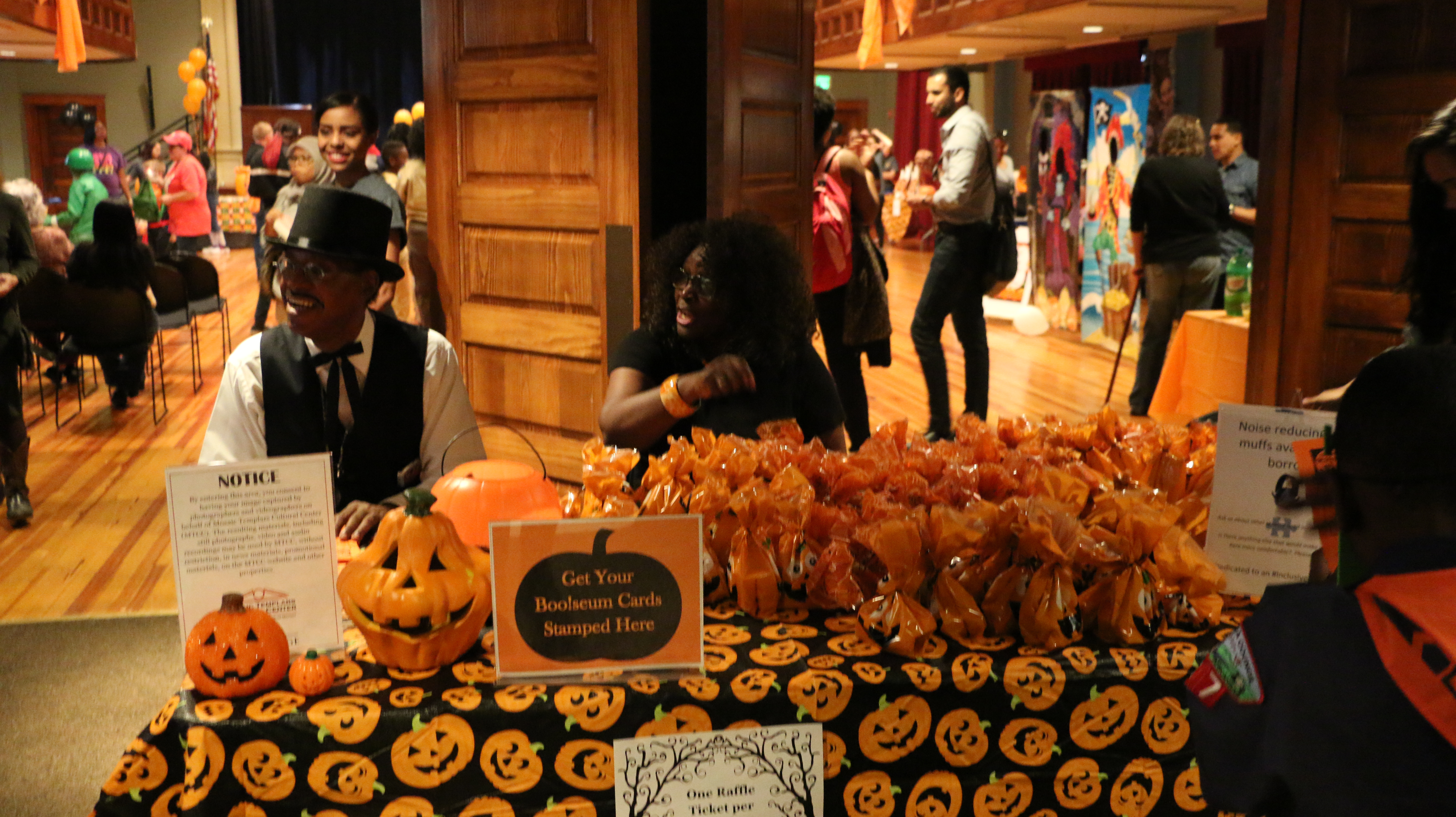 festivalentrancetablewithjack-o-lanterntableclothbucketsandpartyfavorbagsandpeoplesittingbehindit