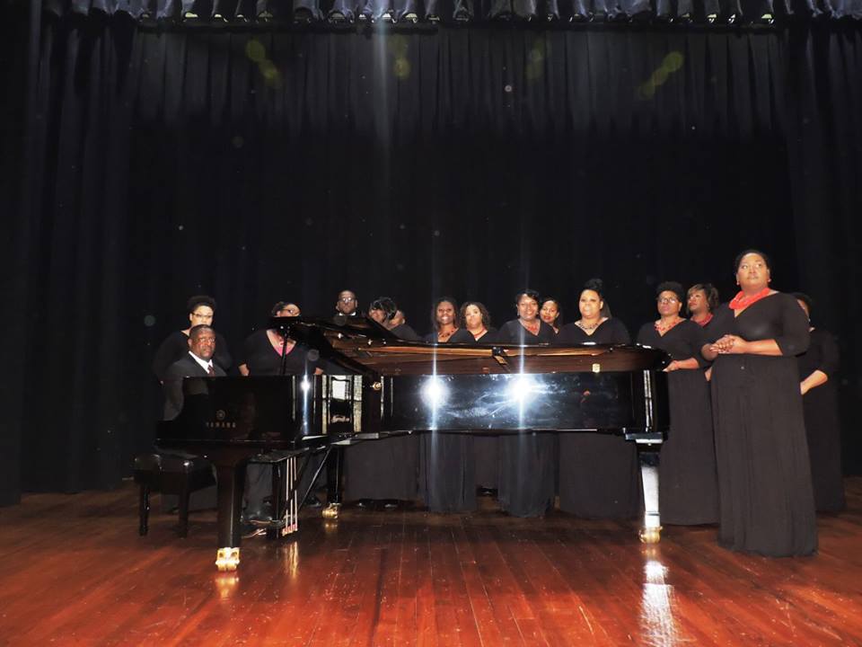 womanschoirwearingblackdressesandrednecklacesstandingaroundpianoandmanplayingitonstage
