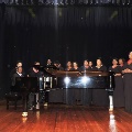 womanschoirwearingblackdressesandrednecklacesstandingaroundpianoandmanplayingitonstage