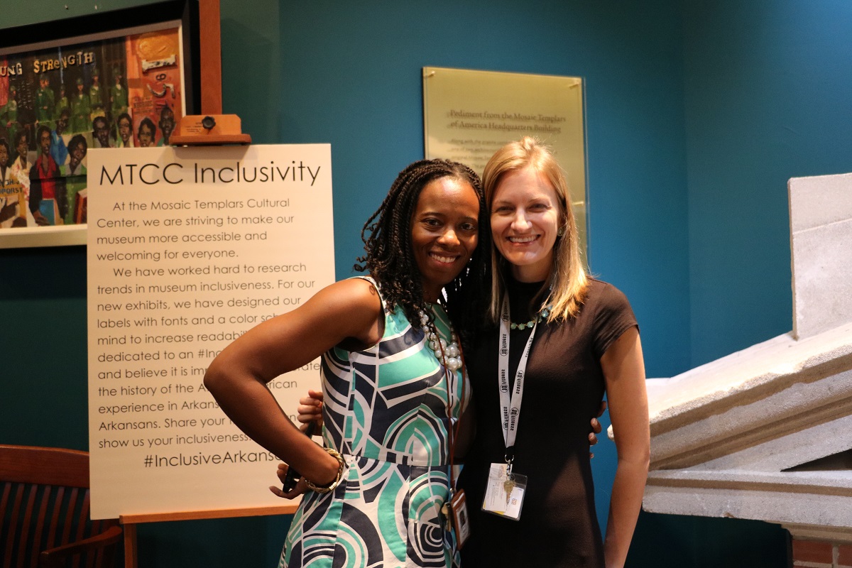 twowomenstandingtogetherandsmilingbyexhibit