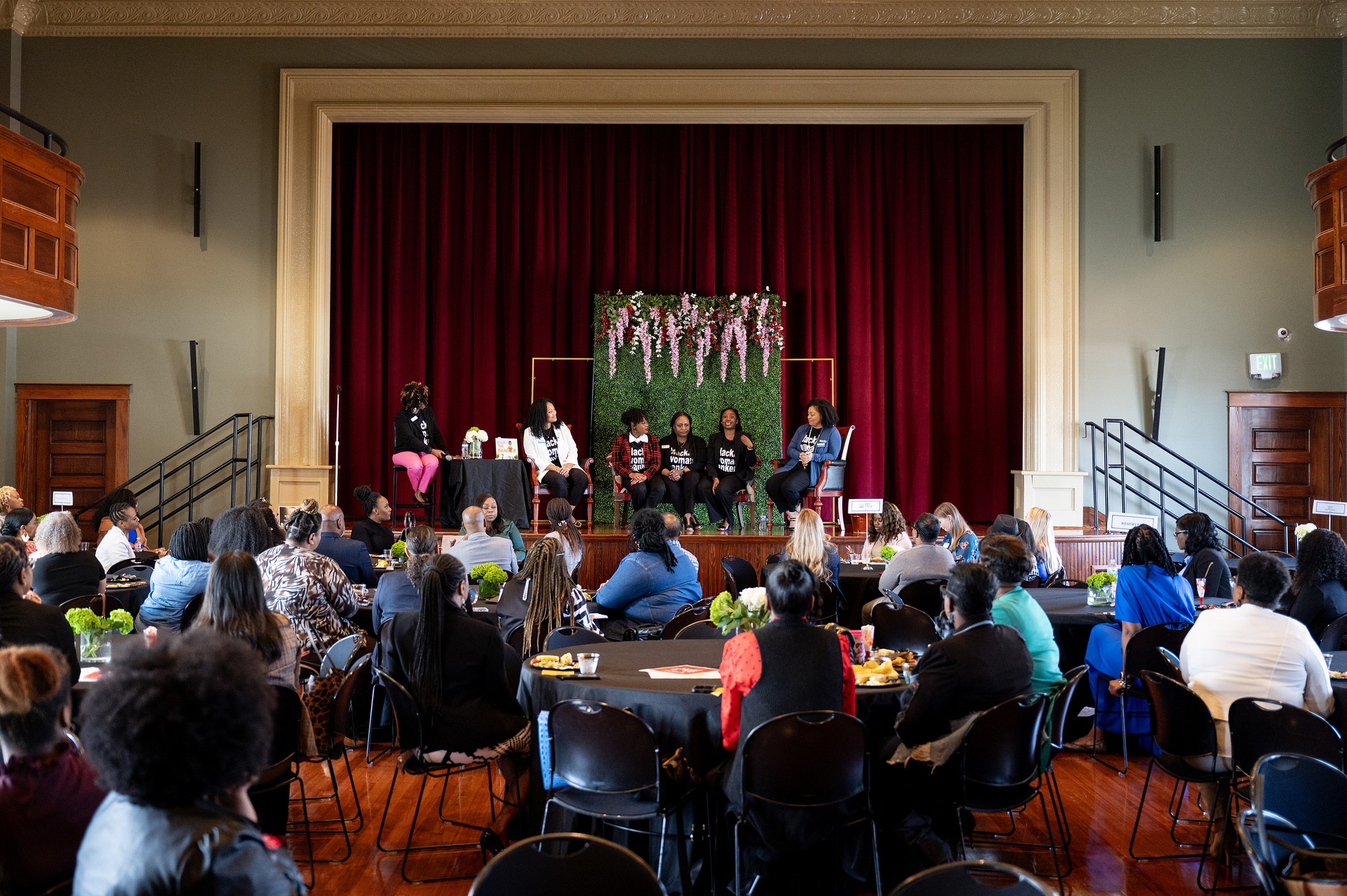 Alargegatheringofpeoplesittingatcirculartableslookingatthestagewhereagroupofwomensitpanel-style